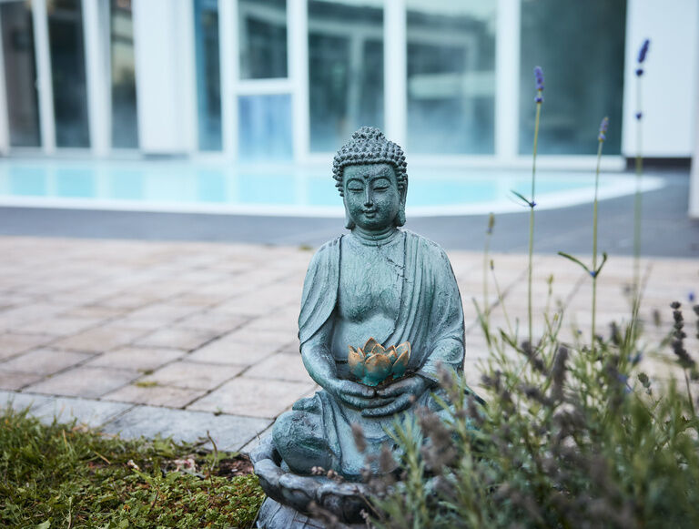 Eine Bronzeskulptur eines sitzenden Buddas vor dem Aussenpool des Thermenhotels Holzapfels in Bayern.