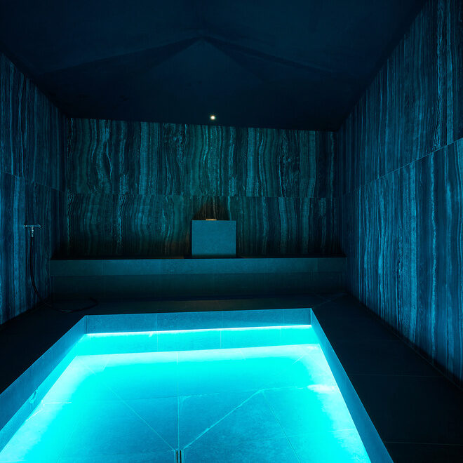 In einem Raum aus dunklen Fliesen ist in der Mitte ein kleiner Pool.