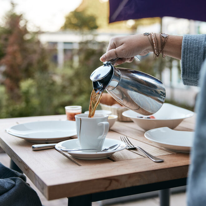 Eine Kaffeetasse wird von einer Person in Bademantel mit frischem Kaffee auf der Terrasse des Thermenhotels Holzapfel in Bayern befüllt.