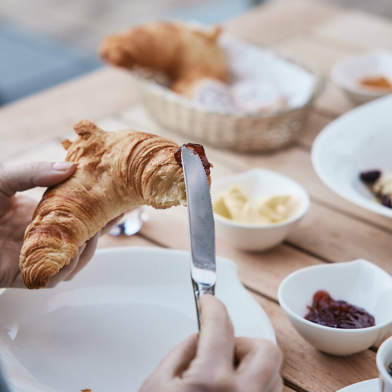 Eine Person trägt  beim frühstück im Hotel Holzapfel in Bad Füssing mit einem Messer Marmelade auf ein Croisant auf.