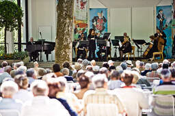 Eine Gruppe von Personen sitzt auf Stühlen in einem Park von Bad Füssing, umgeben von klassischer Musik eines Konzertes.