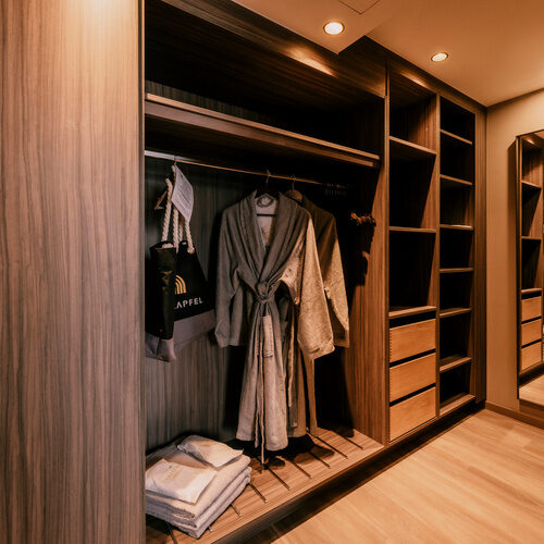 Ein großzügiger begehbarer Kleiderschrank mit Holzoptik und hängender Kleidung darin in der Suite Goldrenette Thermenhotel Holzapfel.