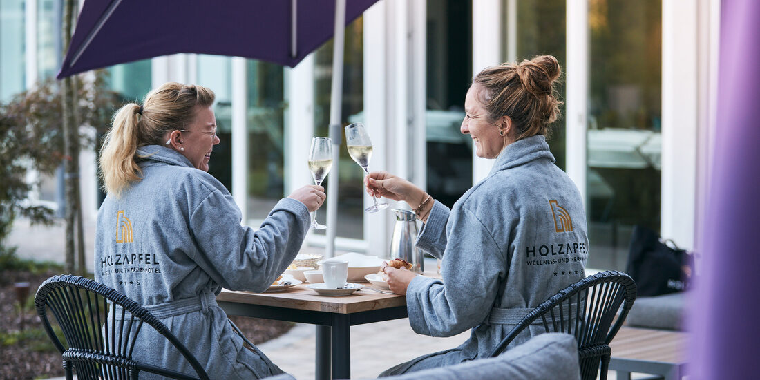 Zwei Frauen sitzen an einem Tisch unter einem Sonnenschirm auf der Poolterrasse des Hotell Holzapfel und stoßen mit einem Glas Wein an.