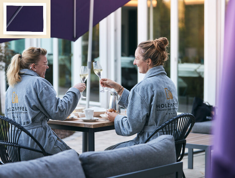 Zwei Frauen sitzen an einem Tisch unter einem Sonnenschirm auf der Poolterrasse des Hotell Holzapfel und stoßen mit einem Glas Wein an.