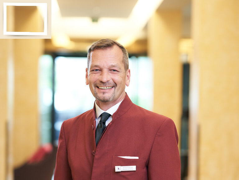 Der Restaurantleiter Roman Sinclair in formeller roter Kleidung, sichtbar zufrieden im Hotelflur des Gesundheitshotels Holzapfel bei Passau.