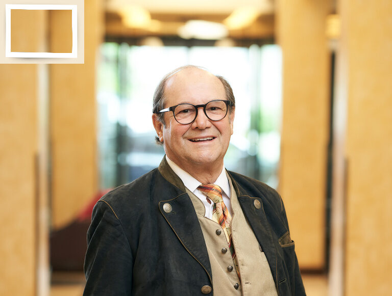 Der Seniorchef Josef Holzapfel steht in Anzug und Krawatte im eleganten Flur des Wellnesshotel Holzapfel in Bad Füssing.