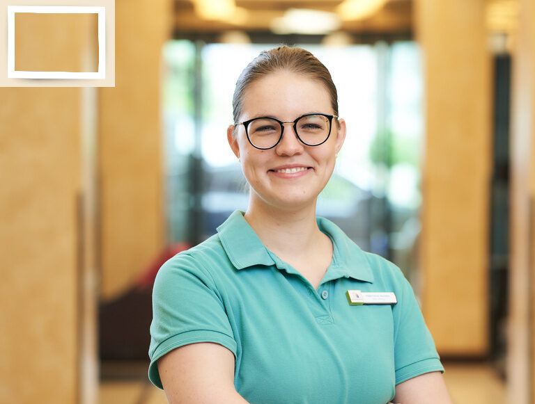 Die Spa Leitung Stephanie Weideck trägt ein grünes Polohemd und blickt positiv vor dem Hoteleingang des Wellnesshotel Holzapfel bei Passau..