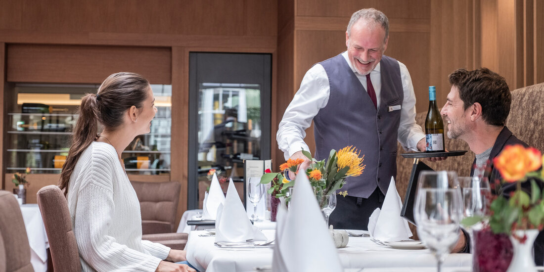 Zwei Personen genießen ein Dinner in der Weinlounge des Hotel Holzapfel im Rottal mit erlesenen ausgeschenkten Weinen von einem Kellner an einem fein gedeckten Tisch.