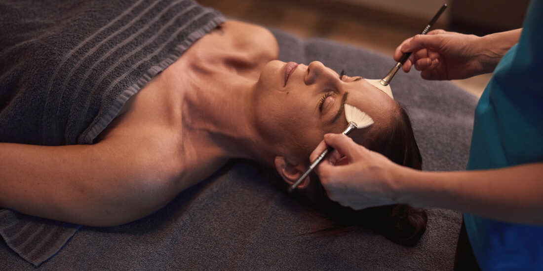 Eine Frau erhält von einer Hotelmitarbeiterin eine entspannende Pinselmassage im Spabereich des Thermenhotels Holzapfel.